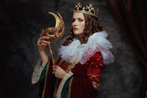 medieval queen in red dress with venetian mask, white collar and crown on dark gray background.