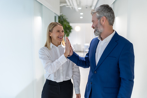 Joyful modern businesspeople giving each other the high five while standing in the office corridor
