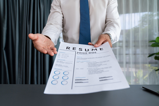 Confident man wearing suit in formal office, hand holding resume paper during job interview. Interviewer point of view with candidate handing resume paper in front of camera for consideration. Fervent