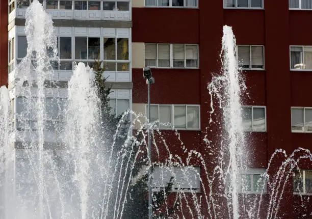 Ornamental water fountain, townsquare, residential buildings exterior in the background, rows of windows. Galicia, Spain.