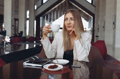 Stylish businesswoman in white elegant suit drinking coffee in cafe while sitting at table and looking away