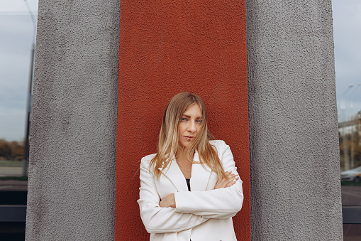 Confident female entrepreneur in white suit and with ambitions standing with crossed arms near wall of building in city and looking at camera