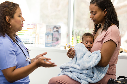 The mature adult doctor gestures as she explains the dosage and side effects of a medication to the young adult mother with her baby.