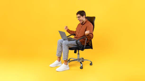 istock Joyful Guy Shaking Fists Using Laptop Sitting Over Yellow Background 1480360969