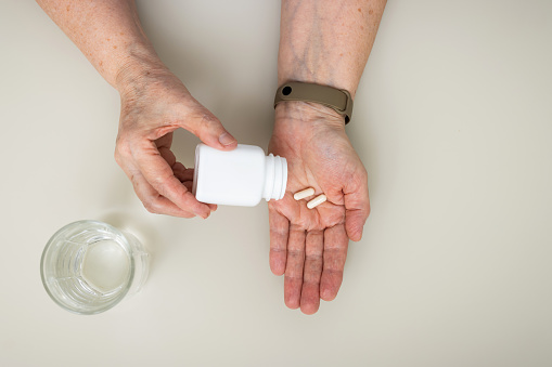Senior woman sitting at the table and pouring from a bottle white capsules, vitamins or pills for treatment into old wrinkled hand, healthcare and medicine concept, top view.