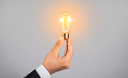 Cropped Shot Of Male Hand In Suit Holding Illuminated Light Bulb, Conceptual Image For Idea And Creativity Concept With Unrecognizable Businessman Standing Over Grey Background, Side View, Copy Space