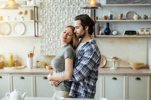 Cheerful happy caucasian millennial hasband with stubble hugs blond wife, enjoy tender moment in light minimalist kitchen interior. Family relationship, love and romance at home, covid-19 quarantine