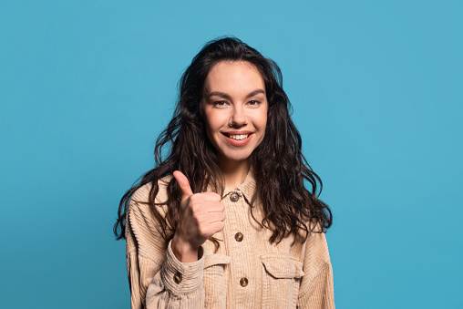 Glad pretty millennial european female student showing thumb up, recommending and advising sale, looking at camera, isolated on blue background, studio, empty space. Emotions, good news, ad and offer