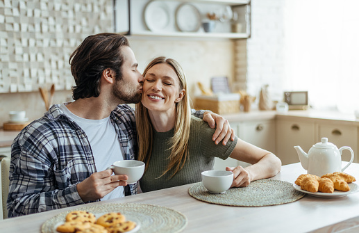 Smiling millennial caucasian husband with stubble hugs and kisses his wife on cheek, couple drinks coffee in modern kitchen interior. Free time, breakfast with drink together and good morning at home