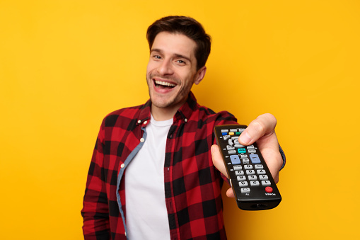 Portrait of excited handsome guy watching TV show or film, holding remote control, switching channels, selective focus on hand. Young smiling man spending weekend free time, yellow studio background