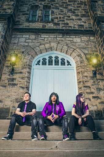 A rock band trio in black and purple sitting on the steps of an old stone building.