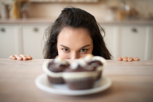 Young woman hiding behind table and looking at delicious cupcakes on plate, focus on lady. Appetite, diet and gluttony concept