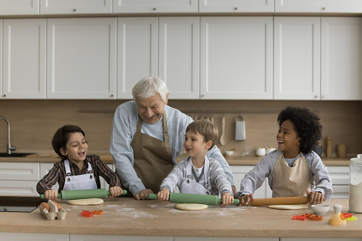Happy grandpa and multiethnic grandsons in aprons preparing cookies, biscuits in kitchen, rolling dough on floury table. Grandfather teaching kids to bake, chatting, smiling, laughing