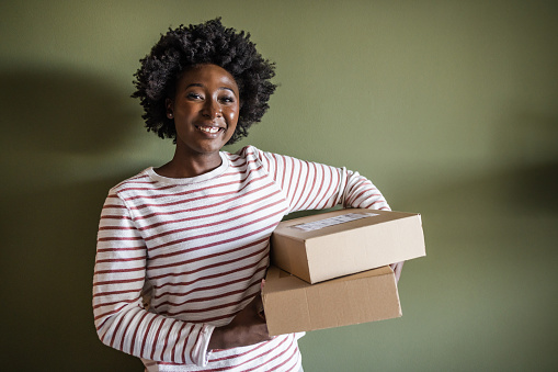 A young  African American woman is standing in front of the wall and holding cardboard boxes. She is smiling and looking at the camera.