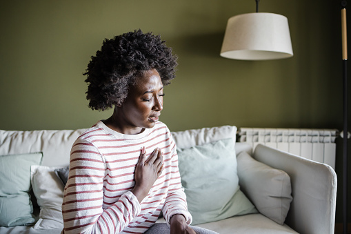 A young African American woman is sitting on the couch at home and holding her chest. Heart disease concept.