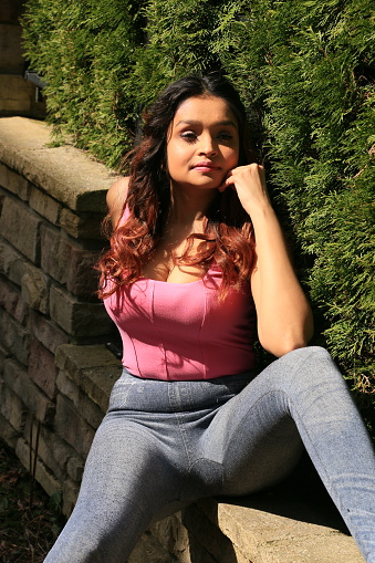 A Fijian model enjoying the Spring sunshine sitting on a rock wall by a hedge. She is wearing long curly brown hair, makeup, a pink sleeveless top and jeans.