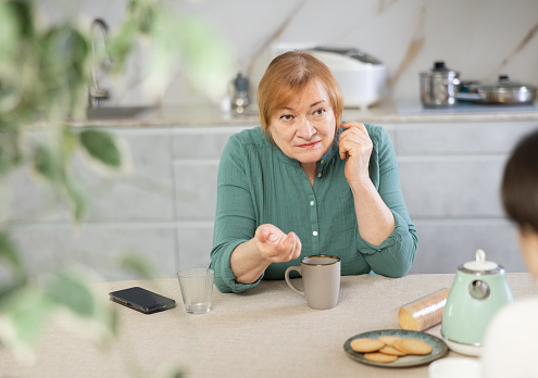 Elderly woman in casual clothes is outraged by communication with her daughter-in-law. Women talk about nasty things in  kitchen at  table and drink tea or coffee