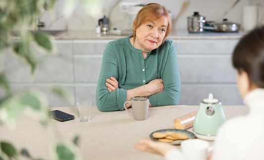 Indignant elderly woman with  cup of tea sits in  kitchen and listens to daughter.Women talk about family difficulties.Concept of troubled family relationships