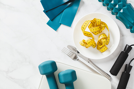 Diet, weight loss concept. Flat lay plate with centimetre tape, cutlery, dumbbells, scales and fitness equipment on white marble background. Top view.
