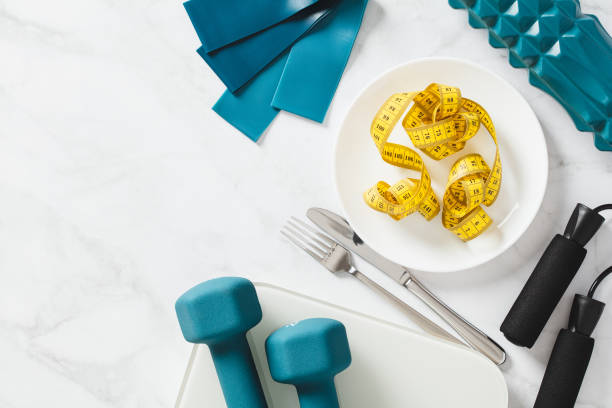 Diet, weight loss concept. Flat lay plate with centimetre tape, cutlery, dumbbells, scales and fitness equipment on white marble background. Top view. Diet, weight loss concept. Flat lay plate with centimetre tape, cutlery, dumbbells, scales and fitness equipment on white marble background. Top view. centimetre stock pictures, royalty-free photos & images
