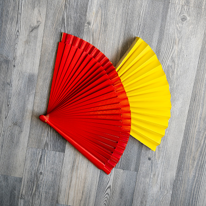 Close up photo of red and yellow colored hand fans on gray colored wooden background. No people are seen in frame. Shot under daylight.