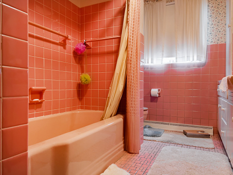 An outdated vintage bathroom highlighted in pink and flowered wallpaper.. The toilet and shower are visible.  The walls are tile squares.  From a middle-class home.