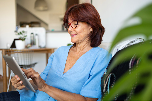 Mature latin Lady with glasses and light blue shirt using digital tablet in the apartment with a lot of plants. High quality photo