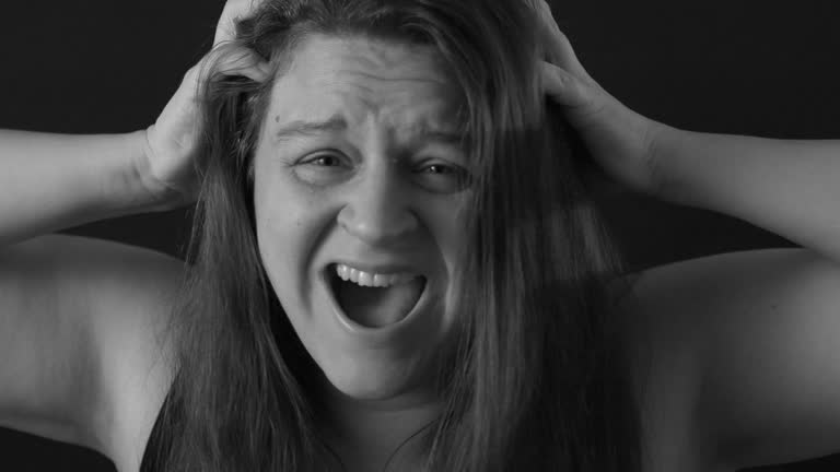 Black and white portrait of crazy terrified middle-aged woman with loose long hair clutching head with hands, screaming.