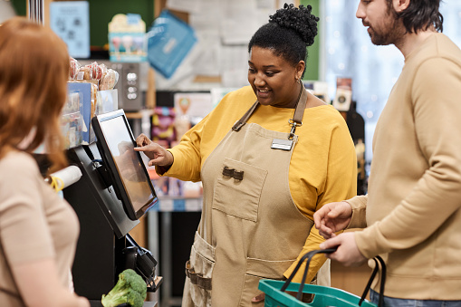 Waist up portrait of smiling black woman helping customers with self checkout in supermarket