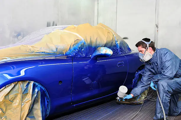 Worker painting a whole side of a blue car in a paint booth.