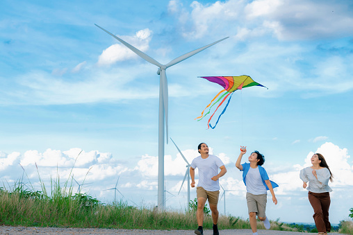 Progressive happy carefree family vacation concept. Young parents mother father and son run along and flying kite together on road with natural scenic on mountain and wind turbine background.