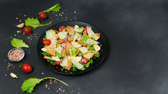 Caesar Salad with chicken, lettuce leaves, cherry tomatoes, grated parmesan in a black plate against a Black Background