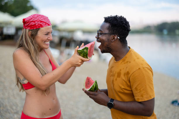 A young multiracial couple near a river is eating watermelons. A young woman and her African man is enjoying watermelon on a warm sunny day near the water. black man eating watermelon stock pictures, royalty-free photos & images