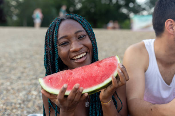 A photo of a young African woman and her male friend with watermelons near the river bank. A photo of a young man near the water, enjoying a fresh watermelon on a sunny day with his female friend of African-American ethnicity. black man eating watermelon stock pictures, royalty-free photos & images