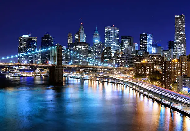 A nighttime view of Manhattan skyline in New York City from the East River strait.  The skyline is in the background, and the strait is in the foreground.  The Brooklyn Bridge is ahead of the skyline, and construction of the One World Trade Center building is visible toward the right side of the scene.