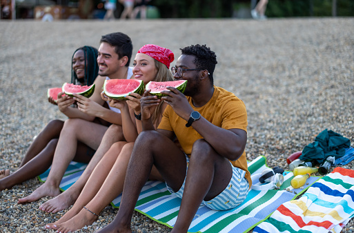 A young multiracial group of friends is enjoying watermelon on a warm sunny day while sitting near the river beach.