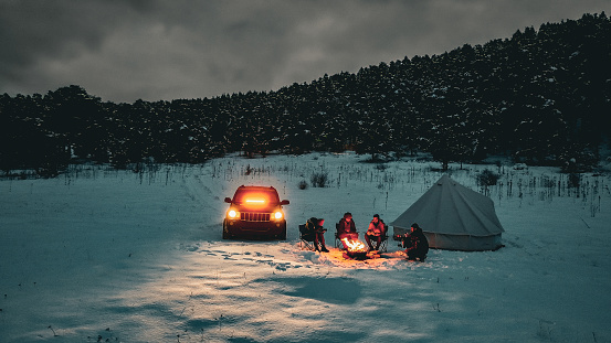 People spending winter holidays in forest, Friends camping on shore of snow-covered winter lake at twilight, camping on snowy mountain, mountain travelers warming up by campfire, aerial view of winter camp