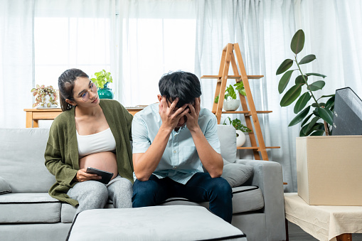 In a living room, Husband put your hands on his face and worry about financial problems because he just dismissed from his work. Wife with near birth sit on sofa nearly and try to encourage him.