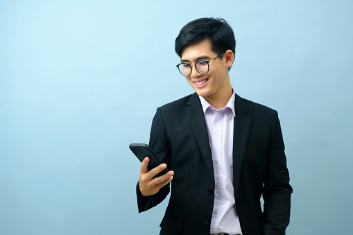 Portrait of young smart asian businessman standing and smiling while using smartphone with light blue isolated background. Business, connection concept.