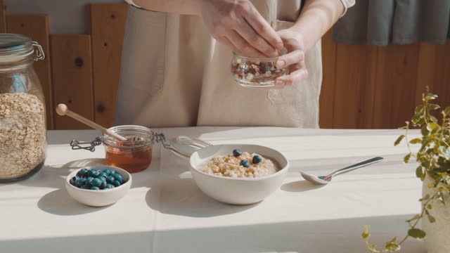Woman preparing healthy dieting vegan nutritious breakfast. Female hand putting blueberries in the bowl with oatmeal porridge with walnuts and honey.