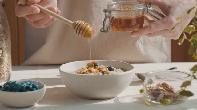 Woman preparing healthy dieting vegan nutritious breakfast. Female hand pouring honey in the bowl with oatmeal porridge with walnuts and blueberries.