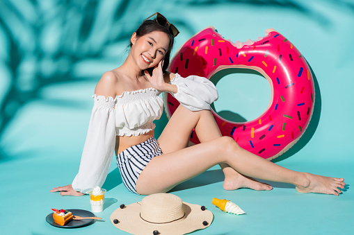 Happy young asia woman slim body wear white and stripe swimsuit sitting relax casual isolated on vivid blue color wall background studio. Summer asian woman fun with inflatable bird and donut summer