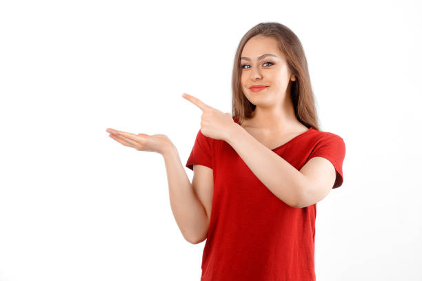 Surprised lovely girl points left at promo banner, shows advertisement on blank white space. Young brunette female looking amazed by promotion offer, stands over white background