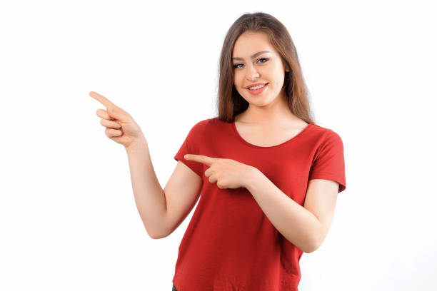 istock Smiling lovely girl points left at promo banner, shows advertisement on blank white space. Young brunette female looking happy by promotion offer, stands over white background 1480553060