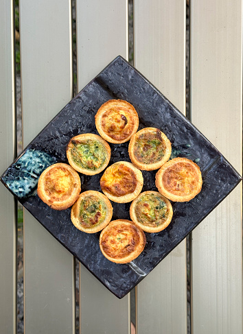 Three Cheese and onion, Spinach Florentine, and Lorraine mini quiche on a plate outdoors.