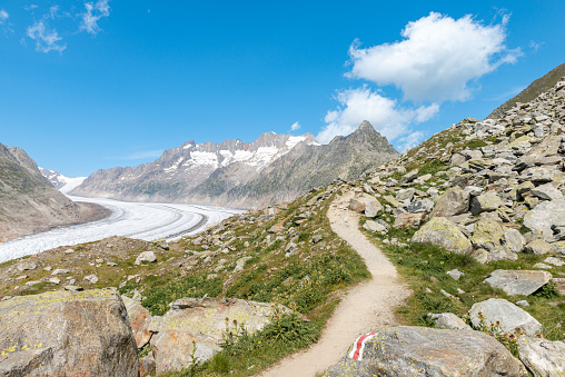 View of a mountain hiking trail with an alpine mountain panorama with the Aletsch Glacier on the left. In the foreground in the middle is the red and white sign of the hiking trail. Near Fiescheralp, Bettmeralp and the Eggishorn in the Jungfrau region, Canton of valais, Switzerland