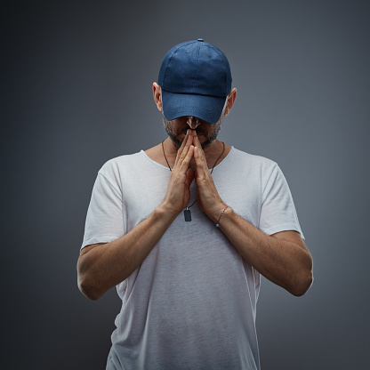 Handsome man wearing white t-shirt and baseball cap, hands folded in prayer against gray background