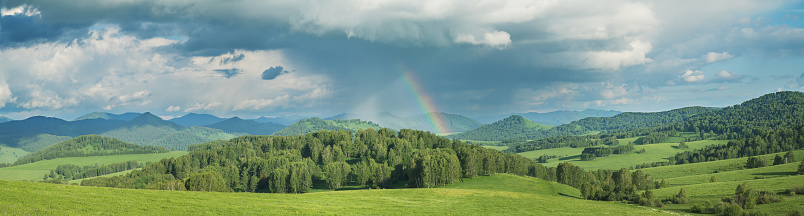 Stormy sky with a rainbow in the countryside, green meadows, mountains and hills