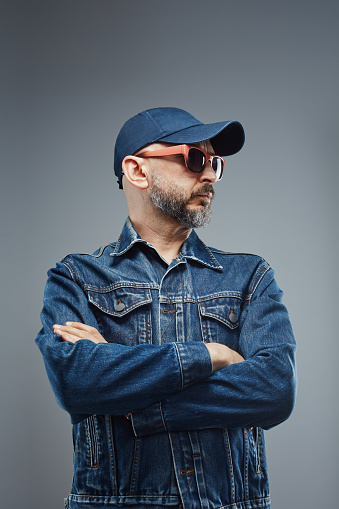 Handsome man wearing denim jacket, baseball hat and colorful sunglasses posing in studio against gray background