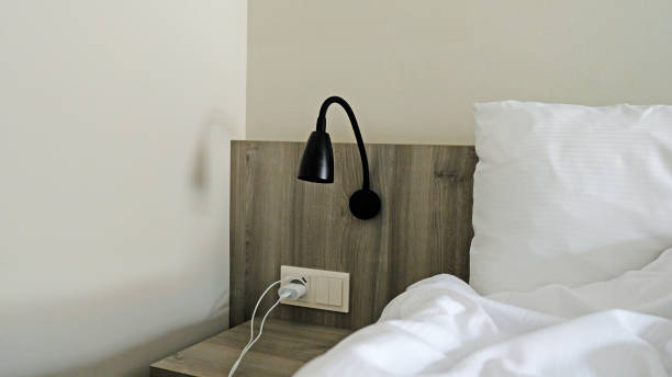 istock Close-up fragment of bedroom with empty bedside table, reading lamp and a USB socket in modern interior​ design home or hotel. Soft pillow and blanket, stylish comfortable furniture. Sun shadows. 1480570228